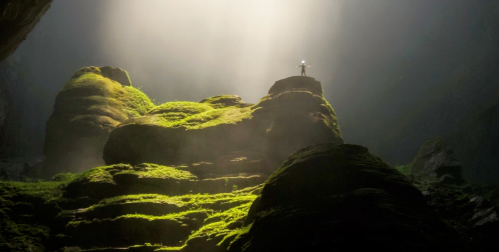 Picture of god rays streaming into a cave, lighting mossy rocks. Someone poses like a starfish at the top of the rock.